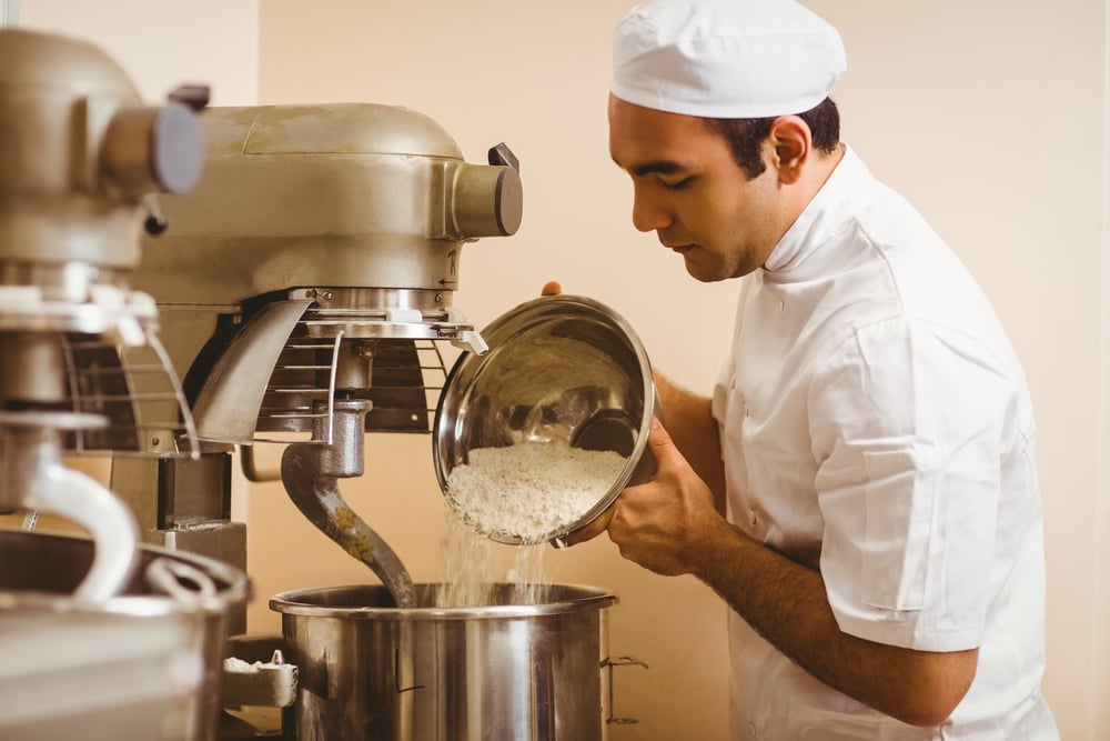 Baker pouring flour into large mixer in a commercial kitchen